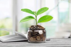 A plant sprouting from a glass jar filled with coins, symbolizing financial growth and investment, placed on a wooden surface next to an open book with a blurred background suggesting a bright, airy space.